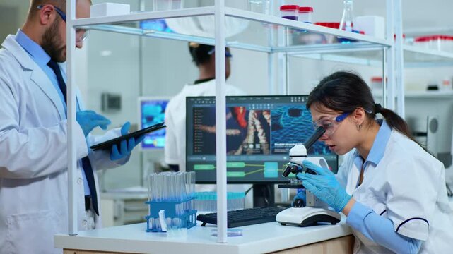 Microscopy, analysis and teamwork as a female researcher studies a specimen while her colleague consults a tablet at a modern laboratory workstation for DNA research, documentation and data review.