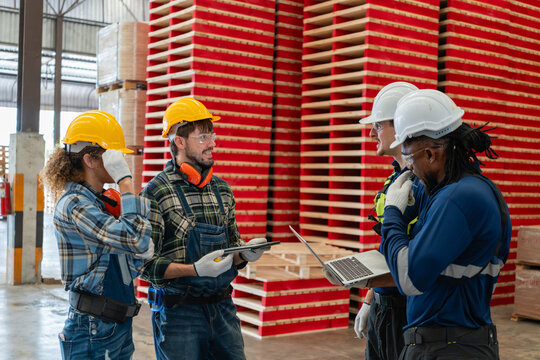 During teamwork discussions, four factory workers hold a team meeting with a laptop and a tablet beside red pallets.