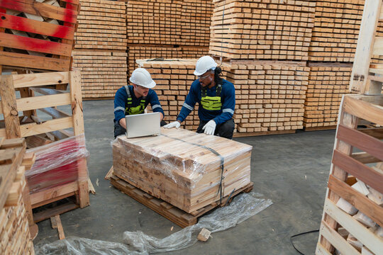 Lumber inventory is checked in the warehouse by two men kneeling with a laptop beside a pallet of wrapped timber.