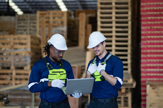 Inside a pallet warehouse, two logistics colleagues consult a laptop together while standing beside wooden pallet stacks.