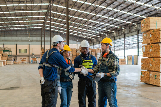 Factory workers show teamwork at a team meeting: two shake hands while two watch, with a participant holding a tablet.