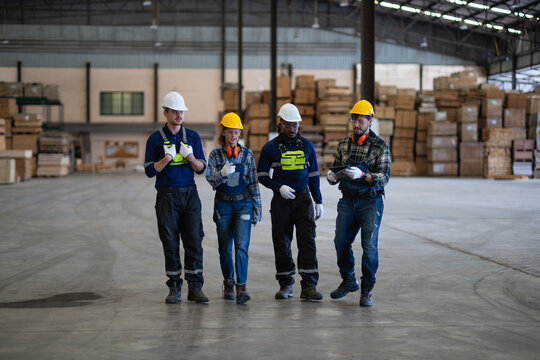 Pallets surround factory workers practicing teamwork as four colleagues hold a walking team meeting and consult a tablet.