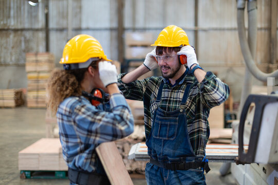 During woodworking with plywood, a carpenter adjusts earmuffs while a coworker holds wood beside the wood workshop panel saw.