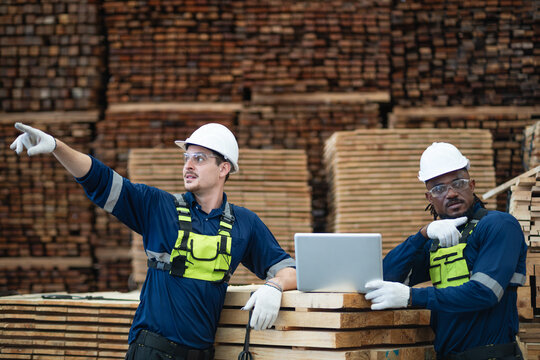 Checking lumber inventory in a warehouse, a man points left while his colleague operates a laptop on stacked timber.