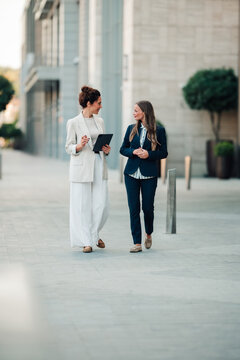 Businesswomen walking and conversing outdoors, collaborating on a project. Focusing on teamwork, communication, and urban professionalism