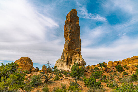A massive, towering sandstone monolith and rock pillar rising from the desert scrub under a wispy blue and white sky.