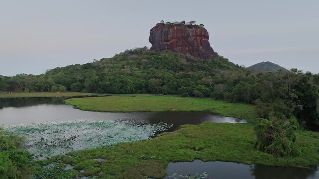 Rock fortress rising above a green wetland and moat at Sigiriya Lion Rock in Sri Lanka, showing grassy foreground and rock formation.