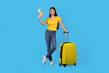 Smiling Asian Woman In Her 20s Holding Travel Documents And Standing Next To Yellow Suitcase, Ready For Vacation. Bright Background Adds Joyful Energy To Travel Theme, Perfect For Adventure Seekers. © Prostock-studio