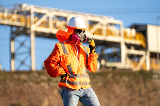 Industrial worker in respirator and high-visibility gear, holding a radio at industrial site