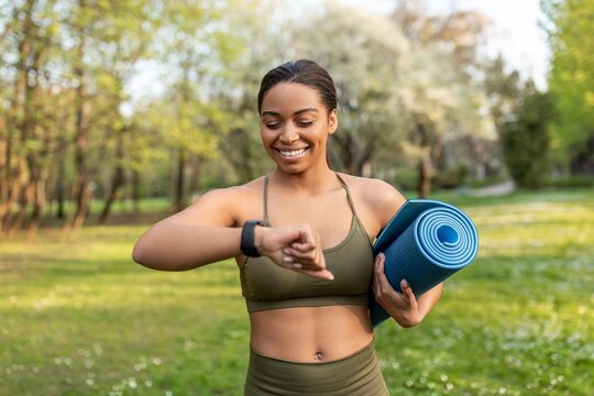 Smiling Black Female In Early Adulthood Checking Fitness Tracker While Holding A Yoga Mat In A Beautiful Park Setting, Enjoying Outdoor Activity And Maintaining Healthy Lifestyle. Weight Loss Concept.