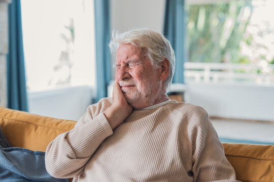 Senior man sitting on a sofa, pressing his hand to his cheek and grimacing from tooth pain, illustrating dental discomfort, aging, and the need for oral healthcare at home