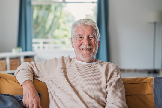 Senior man relaxing on a sofa at home, smiling warmly at the camera in a bright living room and conveying happiness, comfort, wellbeing, and a relaxed retired lifestyle