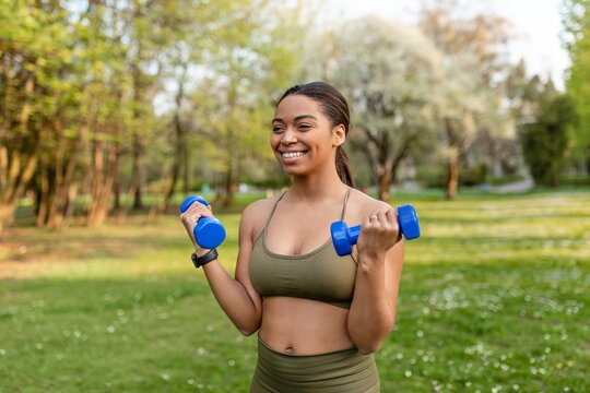 Happy Black Woman In Early Adulthood Exercising With Dumbbells In Peaceful Green Park Setting, Engaging In Fitness Activity To Stay Fit And Healthy, Embracing Outdoor Lifestyle. Weight Loss Concept.