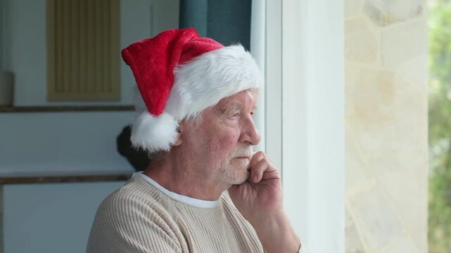 Lonely senior man wearing a red santa hat standing thoughtfully by the window, feeling nostalgic and sad while spending christmas holidays alone at home