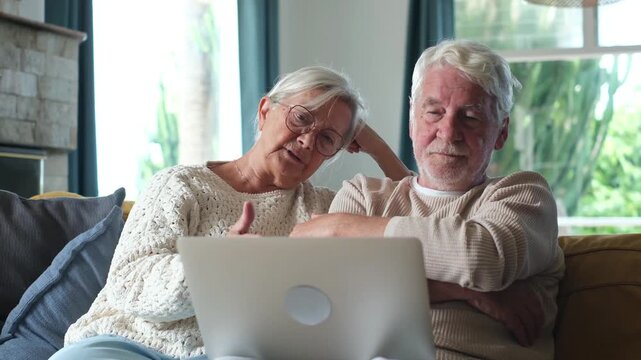 Confused senior couple sitting together on a comfortable sofa, looking at laptop screen with puzzled expressions while trying to understand complicated online information or technology