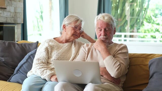 Worried senior couple sitting on a cozy couch at home, looking at a laptop screen with confused and stressed expressions while dealing with online banking issues or bad news