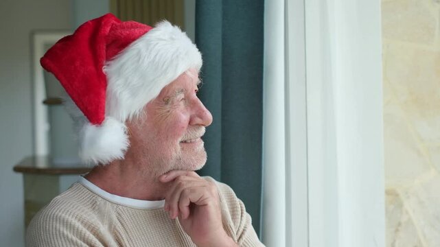 Senior man in a santa hat, smiling thoughtfully with hand on chin while looking through a sunlit window and waiting for family during a cozy christmas holiday at home