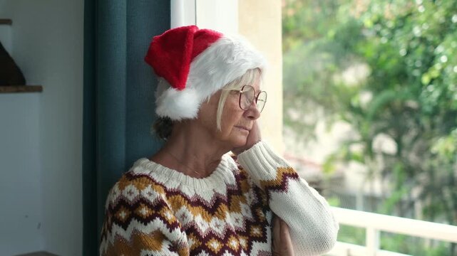 Thoughtful elderly woman wearing a santa hat and eyeglasses standing by the open window, looking outside with a melancholy expression while spending christmas holidays alone at home