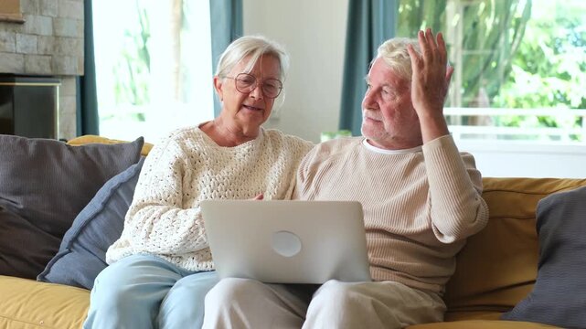 Senior retired couple sitting together on living room sofa, feeling confused and frustrated while dealing with technical computer problems and online financial bills on modern laptop