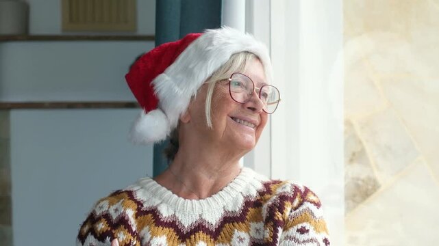 Elderly woman wearing a santa hat and glasses standing near the window, looking outside and smiling thoughtfully while celebrating christmas holiday alone at home
