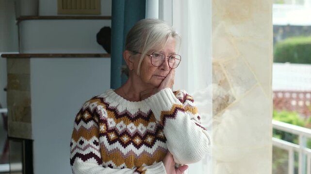 Thoughtful senior woman with eyeglasses resting chin on hand while looking out the window, feeling lonely and melancholic inside a bright modern living room during daytime