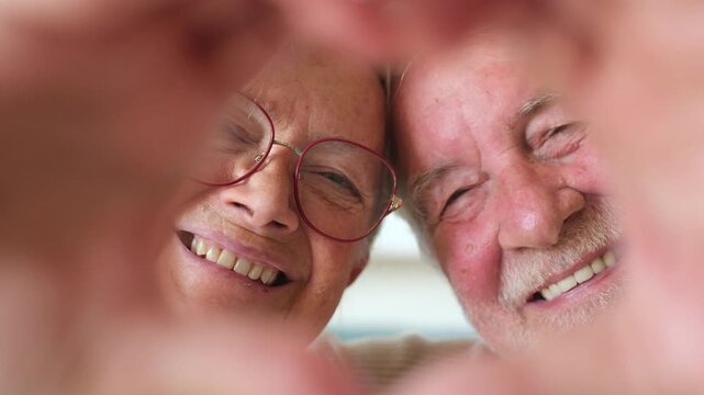 Happy senior couple smiling at the camera through a heart shape created with their hands, expressing enduring love, devotion, romance, and togetherness during retirement years at home