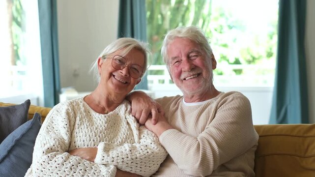 Senior retired couple sitting close together on a comfortable sofa in their living room, looking affectionately at each other before turning to smile warmly at the camera