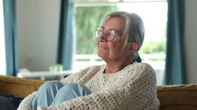 Elderly woman wearing eyeglasses sitting comfortably on a sofa at home, looking out the window while thinking and reminiscing with a serene smile on her face