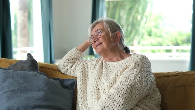 Senior woman with gray hair and glasses relaxing on a cozy living room couch, smiling softly and daydreaming while looking out the window in daylight