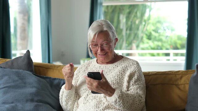 Senior woman sitting on a comfortable sofa at home, receiving good news on her smartphone and celebrating with happy gestures and laughter, enjoying modern mobile technology