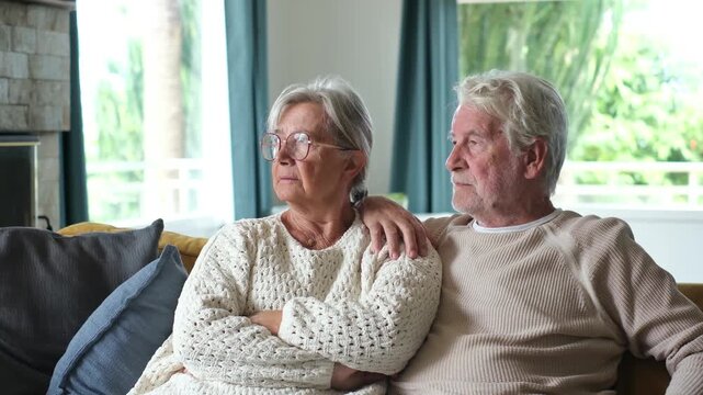Thoughtful elderly husband and wife resting together on a cozy couch at home, looking pensively toward the window while the man gently embraces and comforts his concerned partner