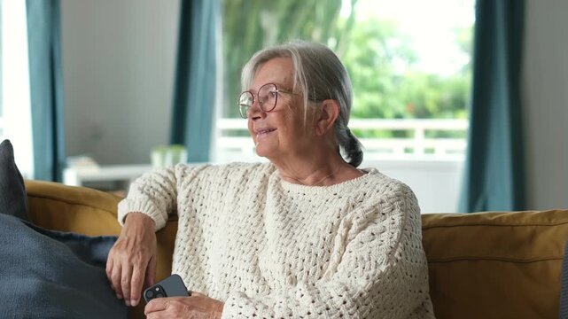 Elderly caucasian woman wearing glasses and knitted sweater sitting relaxed on yellow couch, smiling gently while gazing thoughtfully out the window in her bright living room