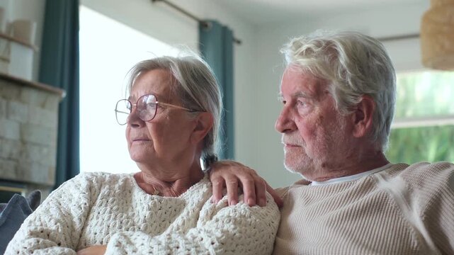 Elderly married couple sitting close together on couch at home, looking out window thoughtfully while elderly husband places comforting hand on wife's shoulder