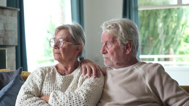 Elderly couple sitting together on couch at home, looking sad, worried, and pensive while husband supports wife by placing arm around her shoulder during difficult moment
