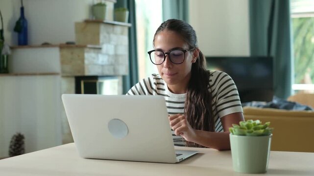 Young businesswoman with eyeglasses typing on laptop keyboard while working remotely at desk in modern living room, smiling and browsing internet content during freelance task