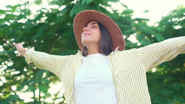Carefree young woman wearing a straw hat standing outdoors in green park, opening her arms wide while smiling and breathing fresh air during sunny summer day