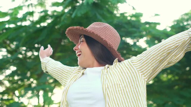 Cheerful young woman wearing a straw hat standing outdoors in green park, spreading her arms wide, inhaling fresh air and enjoying freedom during a sunny summer vacation