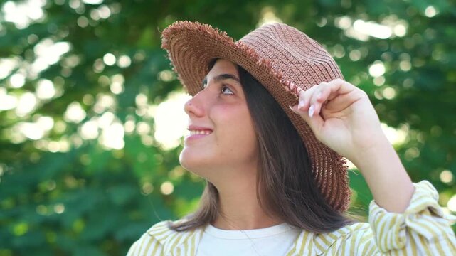 Happy young brunette woman wearing a stylish straw hat smiling, touching headwear, and looking around at green leaves during sunny summer day in park