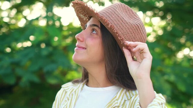 Happy young female tourist wearing a stylish woven sunhat, smiling and looking around while posing amidst lush green foliage in natural golden hour light