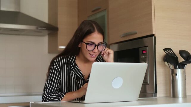 Young female freelancer working on laptop computer in modern kitchen, suddenly celebrating victory and looking happy after receiving great email notification online