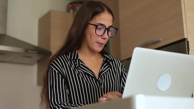 Focused young woman wearing eyeglasses typing on her laptop computer while standing in the kitchen, working remotely as freelancer or studying online from home