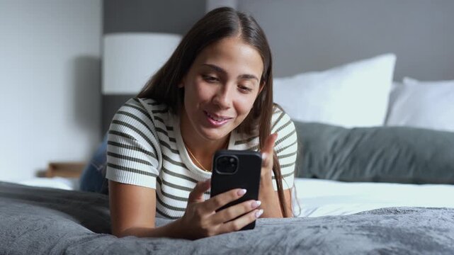 Smiling hispanic woman lying on a comfortable bed in the bedroom, having a cheerful video call with friends using her smartphone while gesturing and relaxing at home