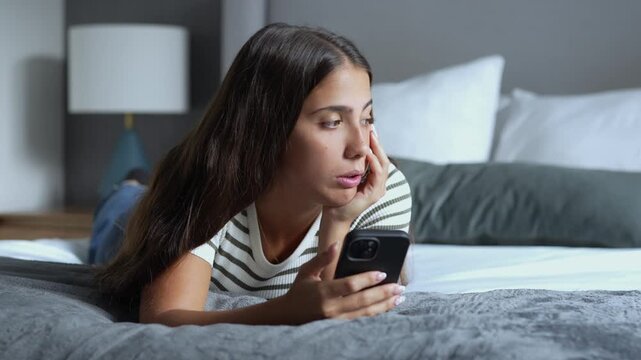 Bored young brunette woman lying on a bed while holding a smartphone, scrolling through social media, sighing with sadness and looking away thoughtfully in a bedroom at home