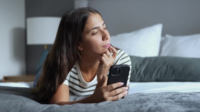 Thoughtful young brunette woman lying on a comfortable bed, holding a smartphone and looking away while pondering and daydreaming in a quiet bedroom in the evening