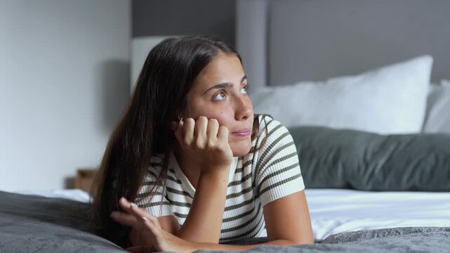 Thoughtful young woman lying prone on comfortable bed, resting chin on hand while looking around pensively and daydreaming inside modern cozy bedroom interior during daytime