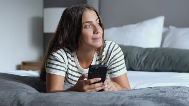Young brunette woman lying comfortably on a bed in a modern bedroom, smiling and thoughtfully daydreaming while typing messages on her smartphone at home
