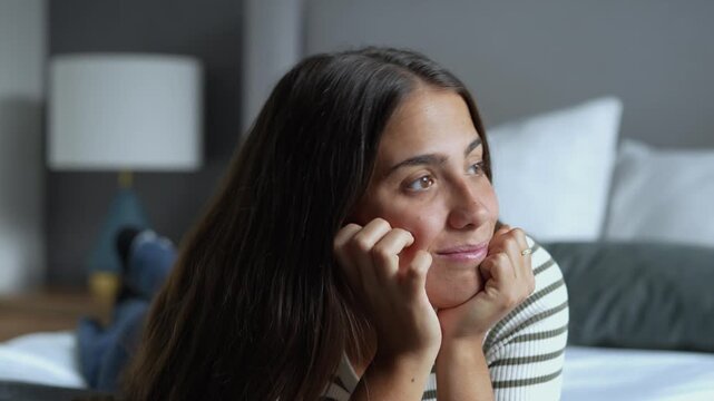 Thoughtful young brunette woman lying comfortably on bed with chin resting in hands, daydreaming and smiling gently while looking away in cozy hotel room