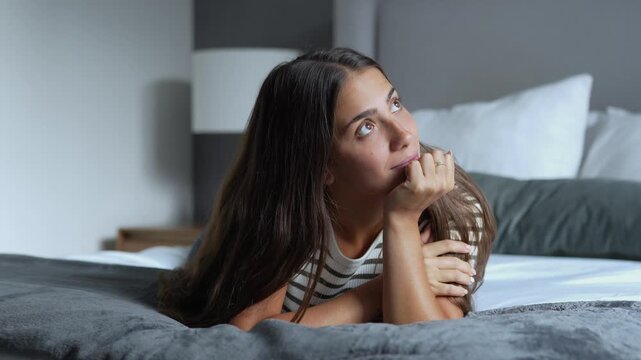 Beautiful young brunette woman lying comfortably on a cozy hotel bed, daydreaming and looking at the camera with a gentle, relaxed smile during morning time