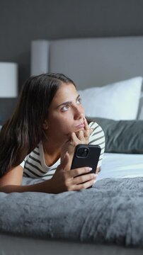 Contemplative young woman lying on bed with smartphone, thinking deeply while waiting for text message or daydreaming indoors during quiet evening relaxation