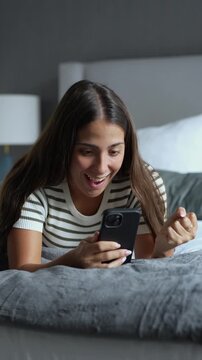 Excited young woman lying on a comfortable bed in the evening, reading good news on her smartphone with a shocked, happy expression and celebrating a sudden online success or victory